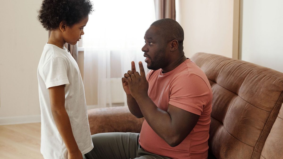 Father sitting on a couch, speaking with his young child who is standing beside him