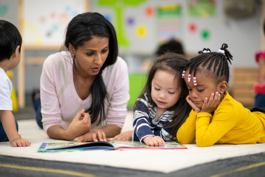 A teacher is lying on the floor with students, one student pointing to a page of the book and asking a question.